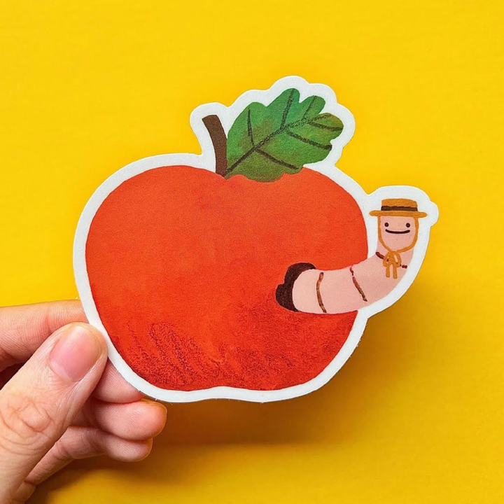 Die-cut sticker of a red apple with a worm and a farmer on a yellow background