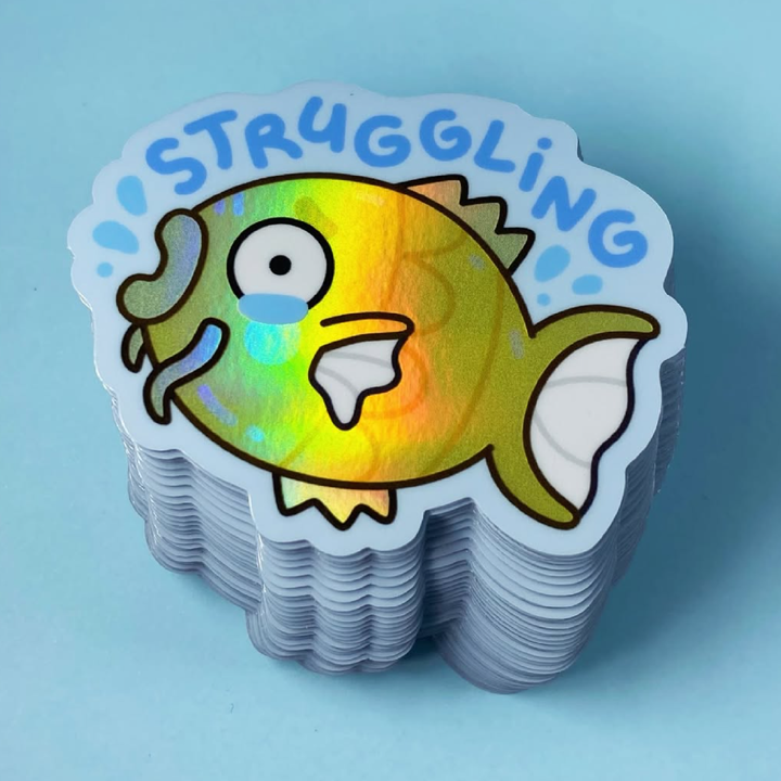 Stack of holographic stickers with a colorful fish design and the word 'STRUGGLING' on a light blue background