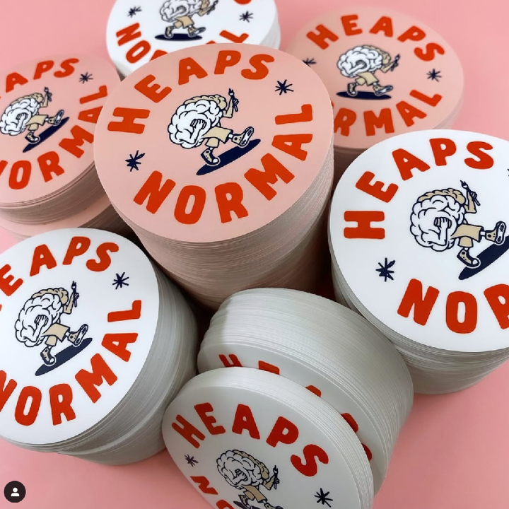 Stacks of circular stickers with 'Heaps Normal' text and cartoon character on a pink background